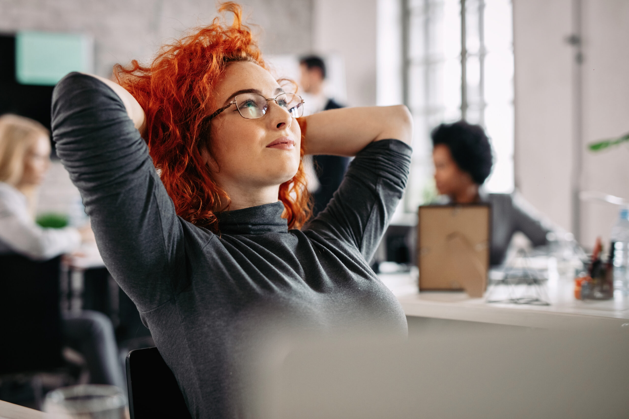 Young pensive businesswoman day dreaming in the office. Satisfied redhead businesswoman resting after work and day dreaming in the office. There are people in the background.